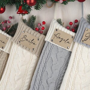 Puede incluir: Cuatro calcetines navideños de punto trenzado en beige, crema y gris, cada uno con una etiqueta de cuero personalizada. Las etiquetas están inscritas con nombres y una huella de pata. La vegetación festiva y las bayas rojas adornan la parte superior.