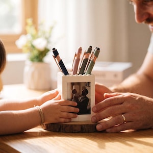 May include: A white pen holder with a photo of a father and child, holding several pens in shades of blue, pink, and green. The pen holder sits on a wooden base. The image shows a father and child.