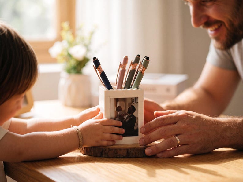 May include: A white pen holder with a photo of a father and child, holding several pens in shades of blue, pink, and green. The pen holder sits on a wooden base. The image shows a father and child.