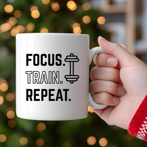 K&ouml;nnte beinhalten: Wei&szlig;e Keramik-Tasse mit dem Text "FOCUS. TRAIN. REPEAT." in Schwarz, zusammen mit einer Hantel-Grafik. Die Tasse hat einen wei&szlig;en Henkel und ist f&uuml;r Fitness-Enthusiasten gedacht.