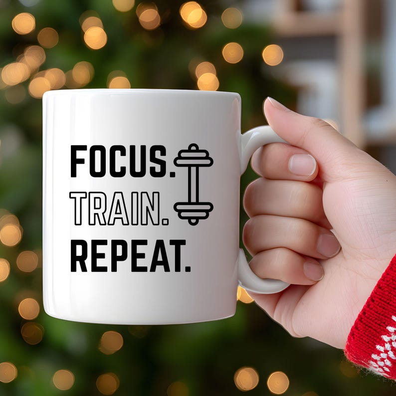 K&ouml;nnte beinhalten: Wei&szlig;e Keramik-Tasse mit dem Text "FOCUS. TRAIN. REPEAT." in Schwarz, zusammen mit einer Hantel-Grafik. Die Tasse hat einen wei&szlig;en Henkel und ist f&uuml;r Fitness-Enthusiasten gedacht.