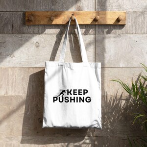 K&ouml;nnte beinhalten: Wei&szlig;e Tragetasche mit schwarzem Text &bdquo;KEEP PUSHING&ldquo; und einer laufenden Figur. Die Tasche h&auml;ngt an einem Holzregal mit Stiften vor einer Holzwand. Das Design ist schlicht und motivierend.