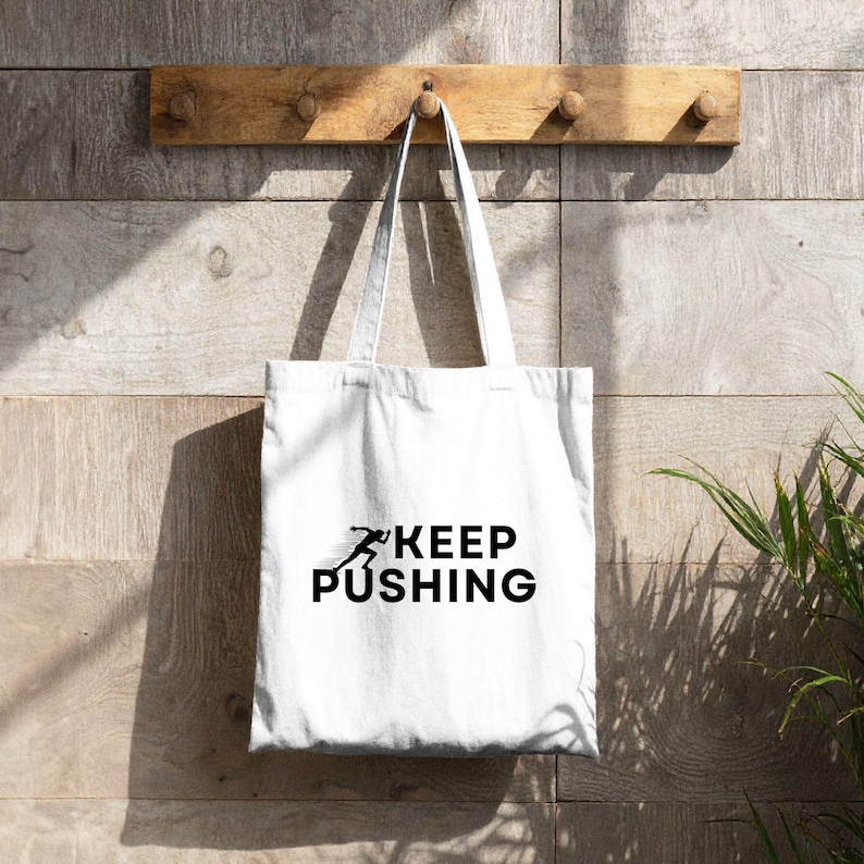 K&ouml;nnte beinhalten: Wei&szlig;e Tragetasche mit schwarzem Text &bdquo;KEEP PUSHING&ldquo; und einer laufenden Figur. Die Tasche h&auml;ngt an einem Holzregal mit Stiften vor einer Holzwand. Das Design ist schlicht und motivierend.