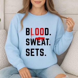 K&ouml;nnte beinhalten: Hellblaues Sweatshirt mit dem Text "BLOOD. SWEAT. SETS." in Schwarz und Rot. Das Wort "BLOOD" ist rot mit einem tropfenden Bluteffekt. Das Sweatshirt hat einen Rundhalsausschnitt.