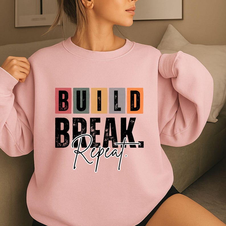 K&ouml;nnte beinhalten: Hellrosa Sweatshirt mit dem Aufdruck "BUILD BREAK Repeat" auf der Vorderseite. Das Wort "BUILD" befindet sich in einzelnen farbigen Bl&ouml;cken. Die W&ouml;rter "BREAK" und "Repeat" sind in schwarzer Schrift.