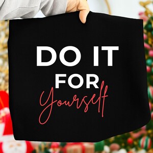 K&ouml;nnte beinhalten: Eine schwarze Tragetasche mit den Worten "DO IT FOR Yourself" in wei&szlig;er und roter Schrift. Die Tasche wird von einer Person hochgehalten. Der Hintergrund ist verschwommen, mit Andeutungen eines Weihnachtsbaums.