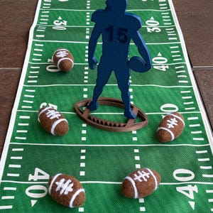 May include: A green football field runner with yard lines and a blue football player figure holding a football. Six brown and white footballs are scattered on the field. The player figure has the number 15 on the jersey.