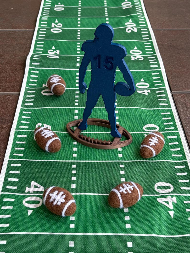 May include: A green football field runner with yard lines and a blue football player figure holding a football. Six brown and white footballs are scattered on the field. The player figure has the number 15 on the jersey.