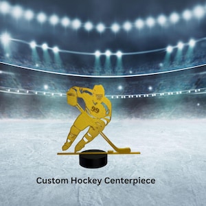 May include: A gold-colored hockey player silhouette with the number 99 on the jersey, holding a hockey stick, and poised on a black puck. The text "Custom Hockey Centerpiece" is displayed below the figure, set against a hockey rink background.