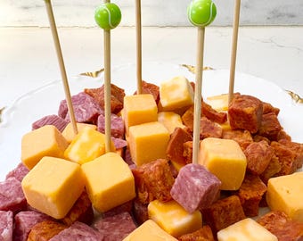 May include: A white plate with an assortment of cubed cheeses and meats, some skewered with decorative tennis ball picks. The cheeses are yellow, white, and marbled, while the meats are red and brown. The picks are green and white.
