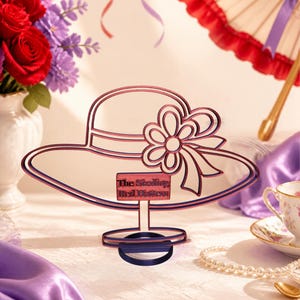 May include: A decorative metal cutout of a wide-brimmed hat with a flower accent on the side. The piece features a small rectangular plaque with the text The Sizzling Red Hatters. The design is finished in a gradient of red and dark blue.