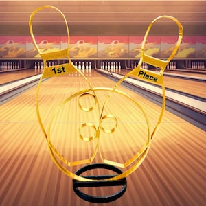 May include: A gold-colored bowling trophy in the shape of two bowling pins with the words "1st Place" on them. The trophy sits on a black base, with a bowling alley in the background.