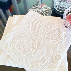 Puede incluir: Servilletas de papel blancas con un dise&ntilde;o floral en relieve. Las servilletas presentan un relieve detallado de grandes flores de peon&iacute;a. Las servilletas est&aacute;n apiladas sobre una superficie oscura, con una copa de vino de cristal y fuentes para servir en el fondo.