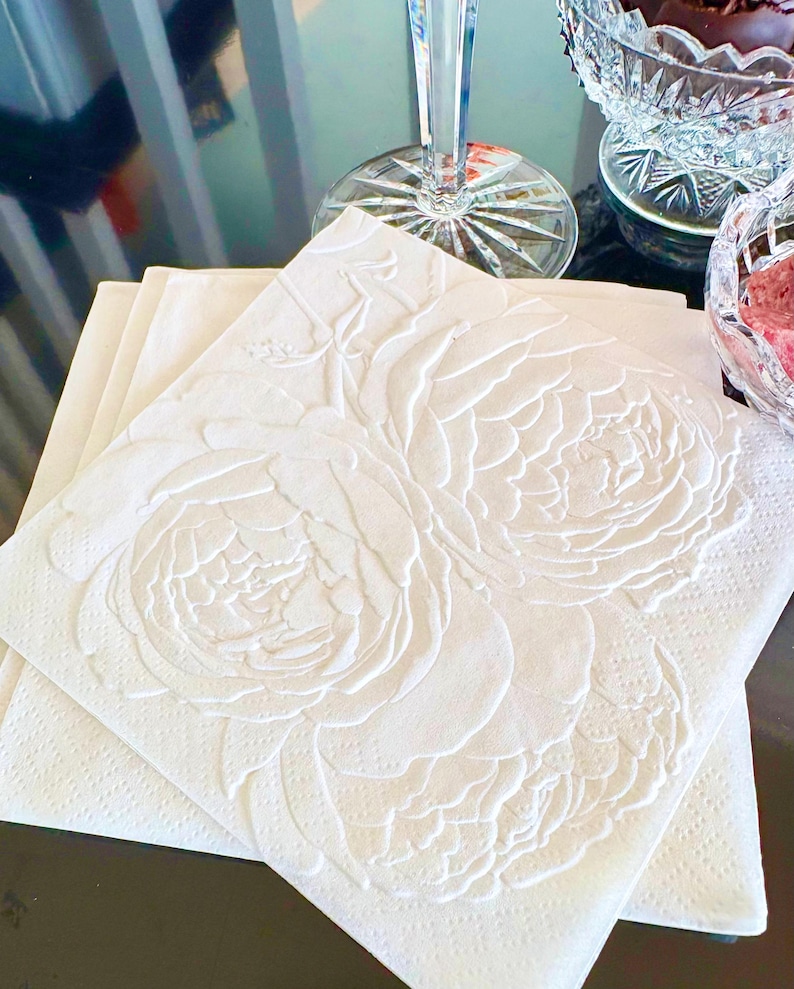 Puede incluir: Servilletas de papel blancas con un dise&ntilde;o floral en relieve. Las servilletas presentan un relieve detallado de grandes flores de peon&iacute;a. Las servilletas est&aacute;n apiladas sobre una superficie oscura, con una copa de vino de cristal y fuentes para servir en el fondo.