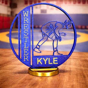 May include: A circular blue 3D-printed trophy featuring a silhouette of a wrestler in a crouched stance. The word WRESTLER is printed vertically on the left side, and the name KYLE is printed at the bottom. The trophy rests on a gold base.