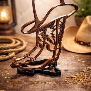 May include: A wooden cowboy boot and hat sculpture. The brown sculpture is intricately cut with a floral design and sits on a black base. A cowboy hat and rope are in the background, suggesting a Western theme.