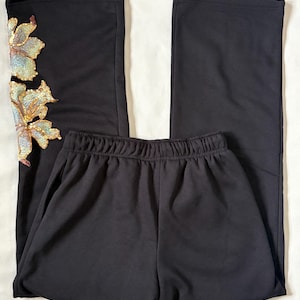 May include: Black pants with a wide leg design. The left leg features an embroidered floral design with gold and iridescent sequins. The pants have an elastic waistband.
