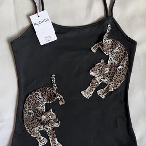 May include: A black camisole featuring two embroidered leopard patches in shades of brown, tan, and black. The garment has thin spaghetti straps and a white rectangular tag attached to the left strap with the text Rhubarb8 printed in black.