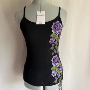 Peut inclure: Un caraco noir à fines bretelles orné d'une broderie florale verticale composée de roses violettes et de feuilles vertes sur le côté droit. Une étiquette blanche attachée à l'encolure porte le texte Rhubarb8 et Tag Us.