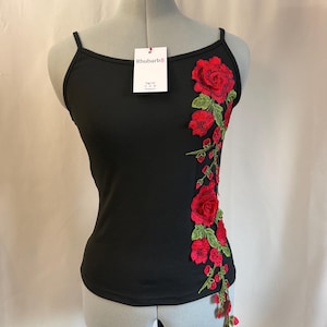 Pode incluir: Uma camisola preta com alças finas e uma aplicação floral vertical de rosas vermelhas com folhas verdes no lado direito. Uma etiqueta retangular branca com o texto Rhubarb8 está presa ao decote.