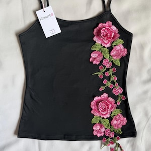 May include: A black camisole with thin shoulder straps featuring a vertical floral embroidery design on the right side. The embroidery consists of pink roses and green leaves. A white rectangular tag with the text Rhubarb8 is attached.