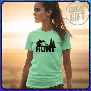 Puede incluir: Camiseta verde claro con una silueta negra de un cazador apuntando a un ciervo, con la palabra "HUNT" debajo. La camiseta tiene mangas cortas y cuello redondo. Un gráfico de "GREAT GIFT" está en la esquina superior derecha.