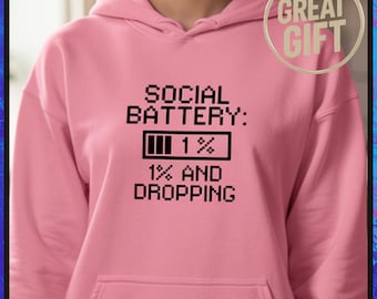 Puede incluir: Sudadera con capucha rosa claro con la frase "SOCIAL BATTERY: 1% 1% AND DROPPING" en negro y un icono de batería baja. Visible una pegatina "GREAT GIFT". La sudadera tiene capucha y bolsillo delantero.