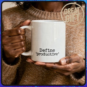 May include: White ceramic mug with the text "Define 'productive'" in black. The mug is held by a person wearing a brown sweater. A gold-colored circle with the words "GREAT GIFT" is in the upper right corner.