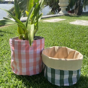 May include: Two fabric plant pots with plaid patterns. One pot is filled with a green plant. The larger pot has a pink and white plaid pattern, while the smaller pot has a green and white plaid pattern with a tan interior.