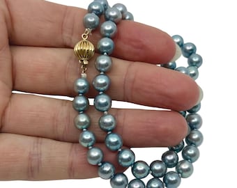 Op de afbeelding: Een ketting met lichtblauwe parels. De parels zijn aan elkaar geregen, waardoor een dubbelstrengs ontwerp ontstaat. De sluiting is goudkleurig en heeft een decoratief, gestructureerd patroon. De ketting wordt in een hand gehouden tegen een witte achtergrond.