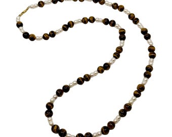Op de afbeelding: Een ketting met afwisselend tijgeroog kralen en kleine, witte parels. De tijgeroog kralen zijn een rijke bruine kleur met gouden strepen. De ketting heeft een goudkleurige sluiting.