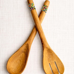 May include: A pair of crossed wooden salad servers featuring a smooth, light brown grain finish. Each handle is decorated with a band of small, multi-colored glass beads in shades of red, yellow, blue, green, and white, arranged in a grid pattern.