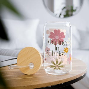 May include: A clear glass can-shaped drinking glass with a bamboo lid and straw. The glass is decorated with pink and peach flowers and the words "Abiding In Christ" and "John 15:4". The glass sits on a wooden table.
