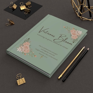 May include: A sage green journal with the words "Virtuous Bloom" and floral accents. The journal includes the quote "She is clothed with strength and dignity, and she laughs without fear of the future." Gold and black pencils and paper clips are also visible.