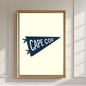 Peut inclure: Impression encadrée avec un fanion bleu marine portant l'inscription "CAPE COD" en blanc. Le fanion est posé sur un fond crème, encadré en or. L'œuvre est exposée sur un mur blanc avec des moulures décoratives.