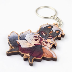 May include: Wooden keychain featuring a cartoon character with a black fox mask, white hair, and a white and red outfit. The character has a grey and white tail.