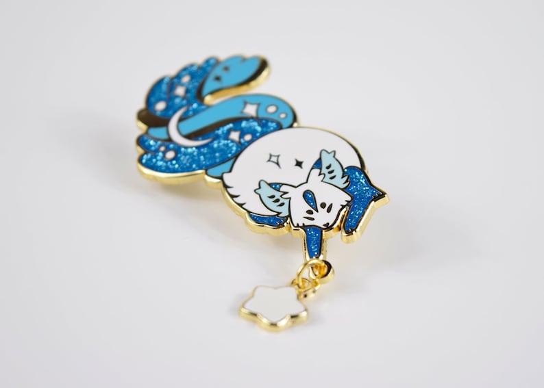 May include: Gold-tone enamel pin featuring a white fox with blue glitter wings and a dangling star charm. The fox is curled up with its tail wrapped around its body.