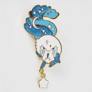 May include: A gold-tone enamel pin featuring a white fox with blue glitter accents. The fox's tail is curled up and has a crescent moon and stars. A dangling star charm hangs from the bottom of the pin.