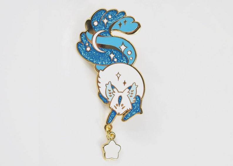May include: A gold-tone enamel pin featuring a white fox with blue glitter accents. The fox's tail is curled up and has a crescent moon and stars. A dangling star charm hangs from the bottom of the pin.