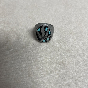 Puede incluir: Anillo plateado con una cara circular negra que presenta un diseño de hoja de cannabis. La hoja está detallada con incrustaciones de color turquesa. El anillo tiene una banda sencilla y clásica.