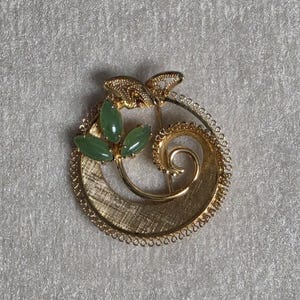 May include: A gold-tone circular brooch featuring a textured crescent base and a delicate wire-work border. Three smooth, oval-shaped green stones are arranged like leaves on a curved stem that spirals towards the centre of the open circle.