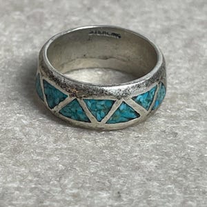 May include: A sterling silver ring with a band of turquoise-colored triangular inlays. The ring has a wide band and a slightly worn appearance. The word "STERLING" is stamped inside the ring.