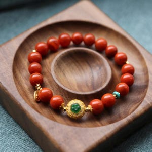 Puede incluir: Una pulsera de cuentas rojas vibrantes, con espaciadores dorados y un dije central dorado con una piedra verde. Una peque&ntilde;a cuenta turquesa a&ntilde;ade un toque de color. La pulsera se muestra en una bandeja de madera.