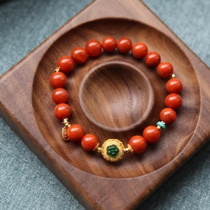 Puede incluir: Un brazalete de cuentas con cuentas grandes y redondas de color naranja. El brazalete incluye detalles dorados y un elemento decorativo central con una piedra verde. Tambi&eacute;n se ve una peque&ntilde;a cuenta turquesa. El brazalete se exhibe en una bandeja de madera.