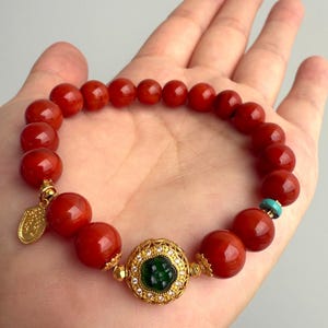 Puede incluir: Una pulsera de cuentas rojas con detalles dorados y una piedra verde central. La pulsera presenta grandes cuentas rojas redondas, un dije dorado y un engaste dorado decorativo alrededor de la piedra verde. Tambi&eacute;n hay una peque&ntilde;a cuenta turquesa.