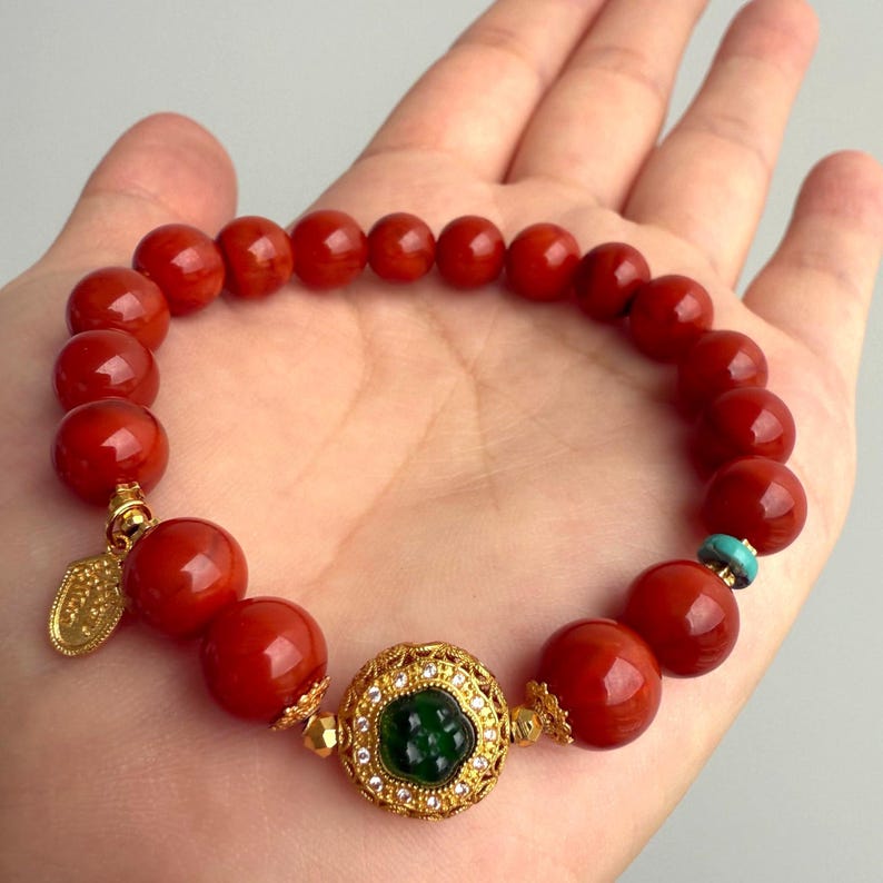 Puede incluir: Una pulsera de cuentas rojas con detalles dorados y una piedra verde central. La pulsera presenta grandes cuentas rojas redondas, un dije dorado y un engaste dorado decorativo alrededor de la piedra verde. Tambi&eacute;n hay una peque&ntilde;a cuenta turquesa.