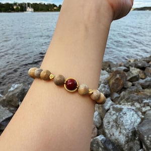 May include: A beaded bracelet featuring light brown wooden beads and a central dark red bead set in a gold-colored frame. The bracelet is accented with small gold-colored spacers. The bracelet is shown on a person's arm.