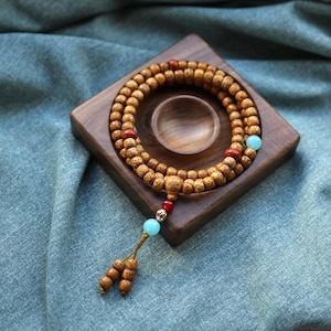 May include: A wooden mala bead necklace with a tassel and turquoise and red accents rests on a square wooden tray. The beads are light brown with a dotted texture. The tray has a circular indentation in the center.