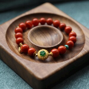 Puede incluir: Una pulsera de cuentas rojas con detalles dorados y una pieza central de piedra verde. La pulsera se exhibe en una bandeja de madera con un dise&ntilde;o tallado. Tambi&eacute;n se ve una peque&ntilde;a cuenta turquesa.