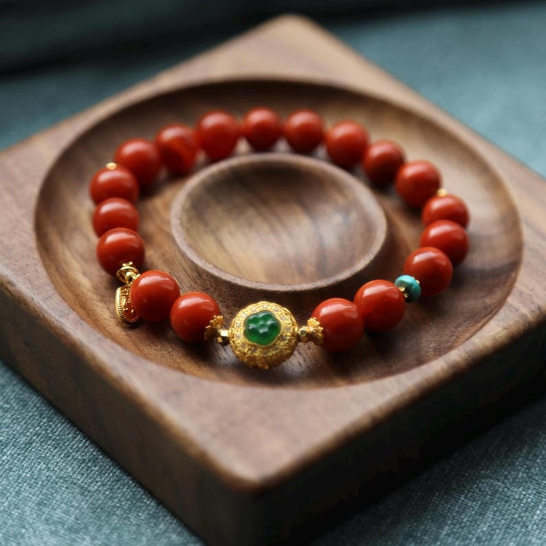 Puede incluir: Una pulsera de cuentas rojas con detalles dorados y una pieza central de piedra verde. La pulsera se exhibe en una bandeja de madera con un dise&ntilde;o tallado. Tambi&eacute;n se ve una peque&ntilde;a cuenta turquesa.